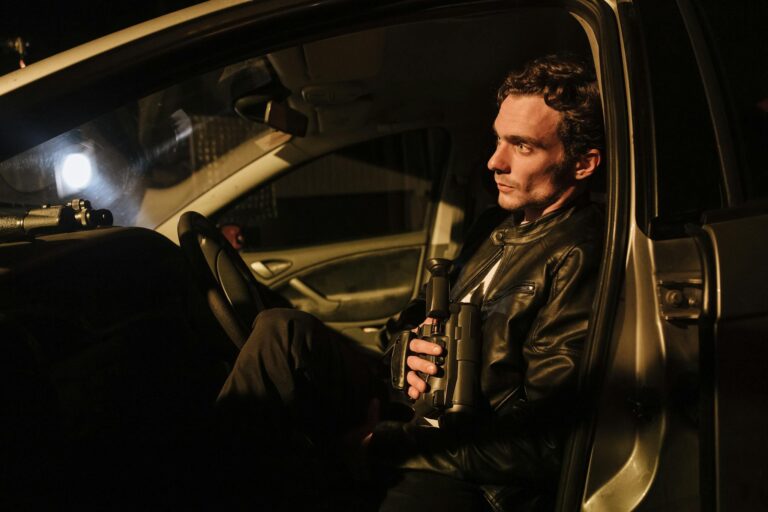 A man sitting in a car holding binoculars, conducting night surveillance.